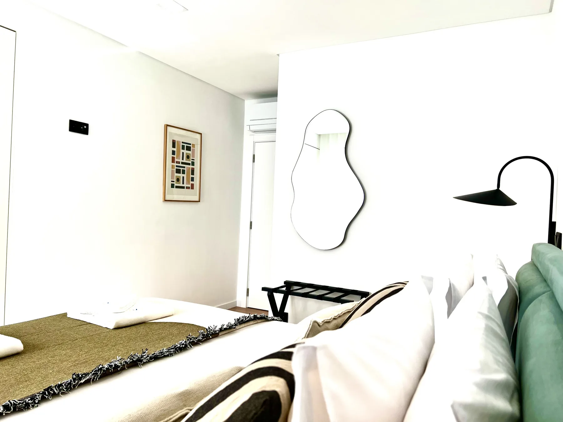 Un dormitorio luminoso con una cama tendida, una lámina enmarcada y una lámpara de pared.