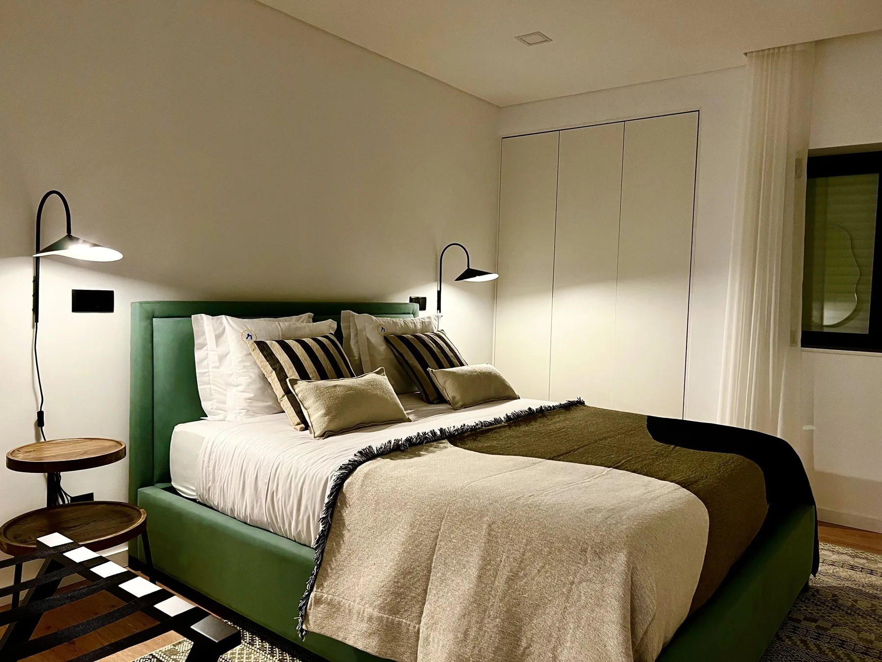 Dormitorio con una cama verde, ropa de cama en capas y luces de lectura en la pared.