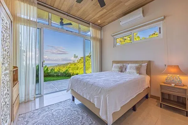 Dormitorio con una cama arreglada junto a grandes ventanas con vistas a vegetación.