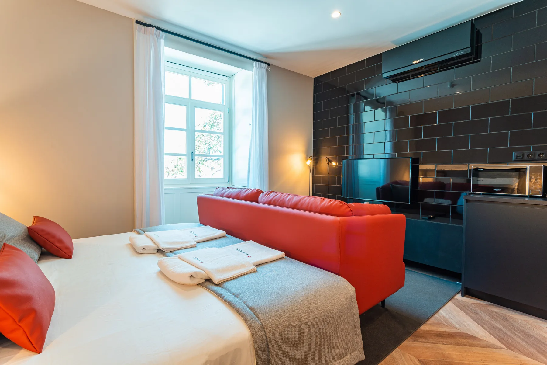 A studio living area with a bed, red seating, and a dark tiled kitchenette.