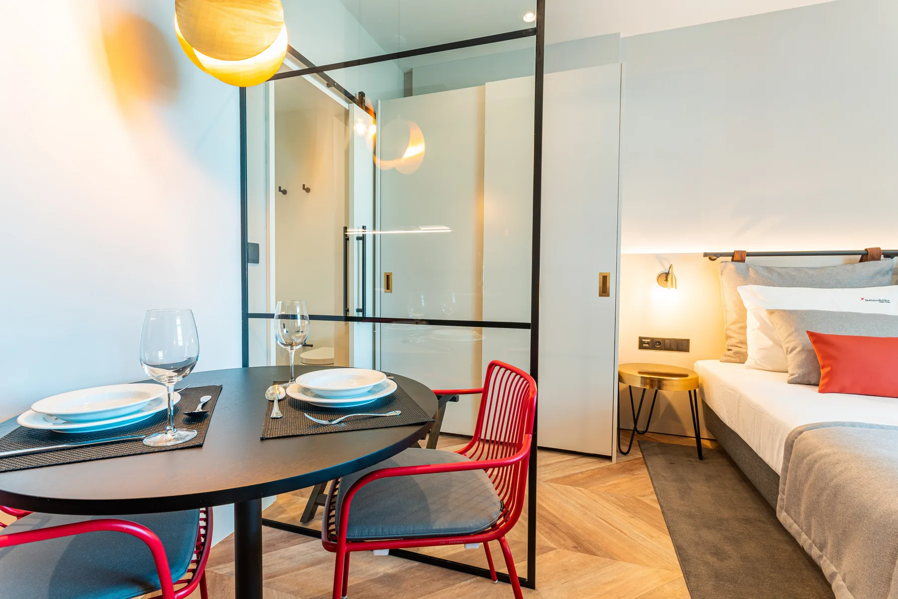 A studio room with a dining table and a bed beyond a glass partition.