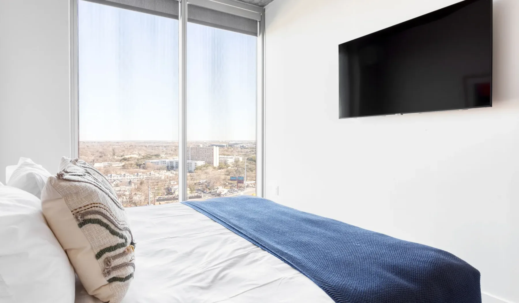 A bedroom with a bed beside a large window and a wall-mounted television.