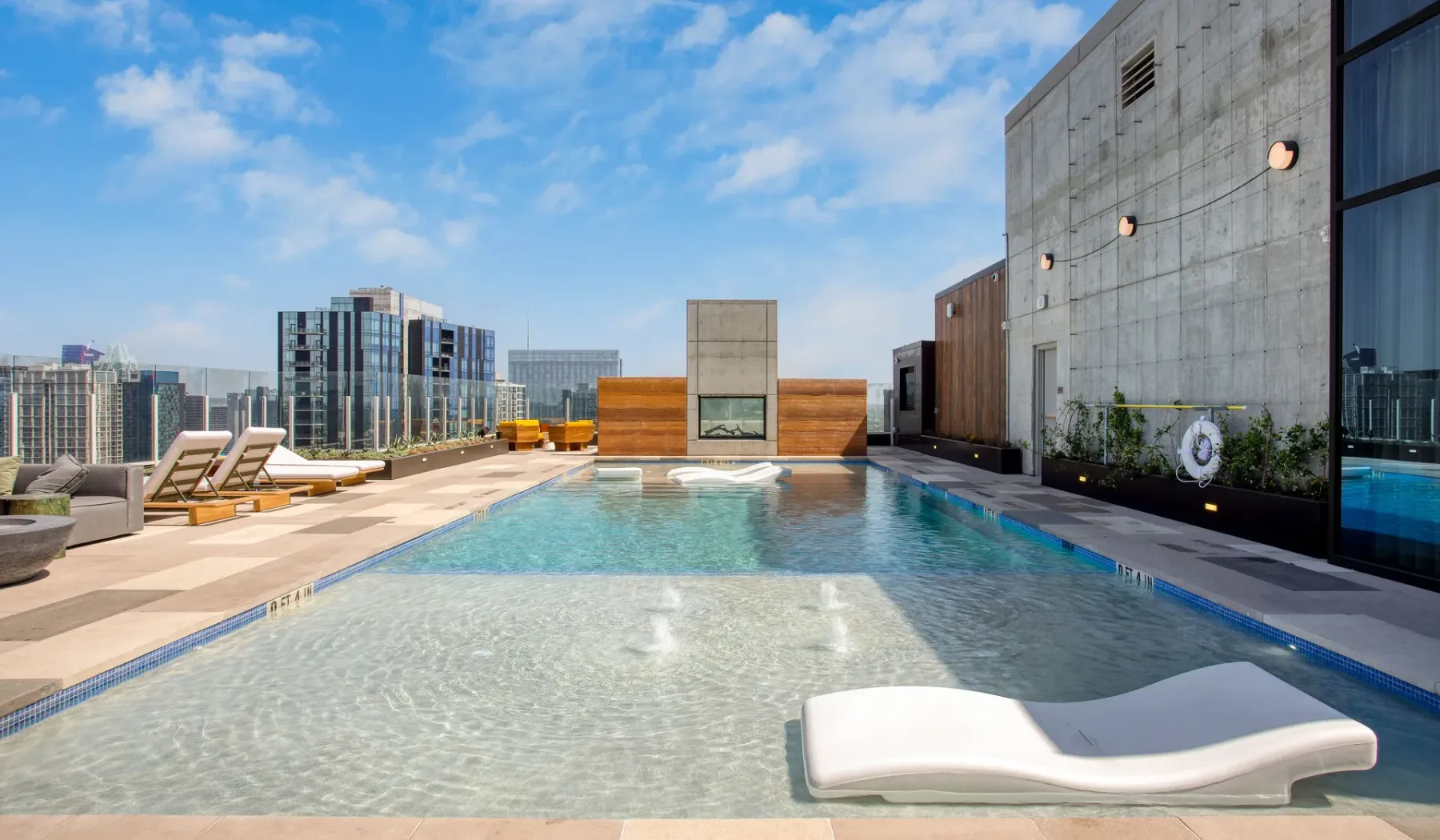 A rooftop swimming pool has a shallow lounging area and city buildings beyond.
