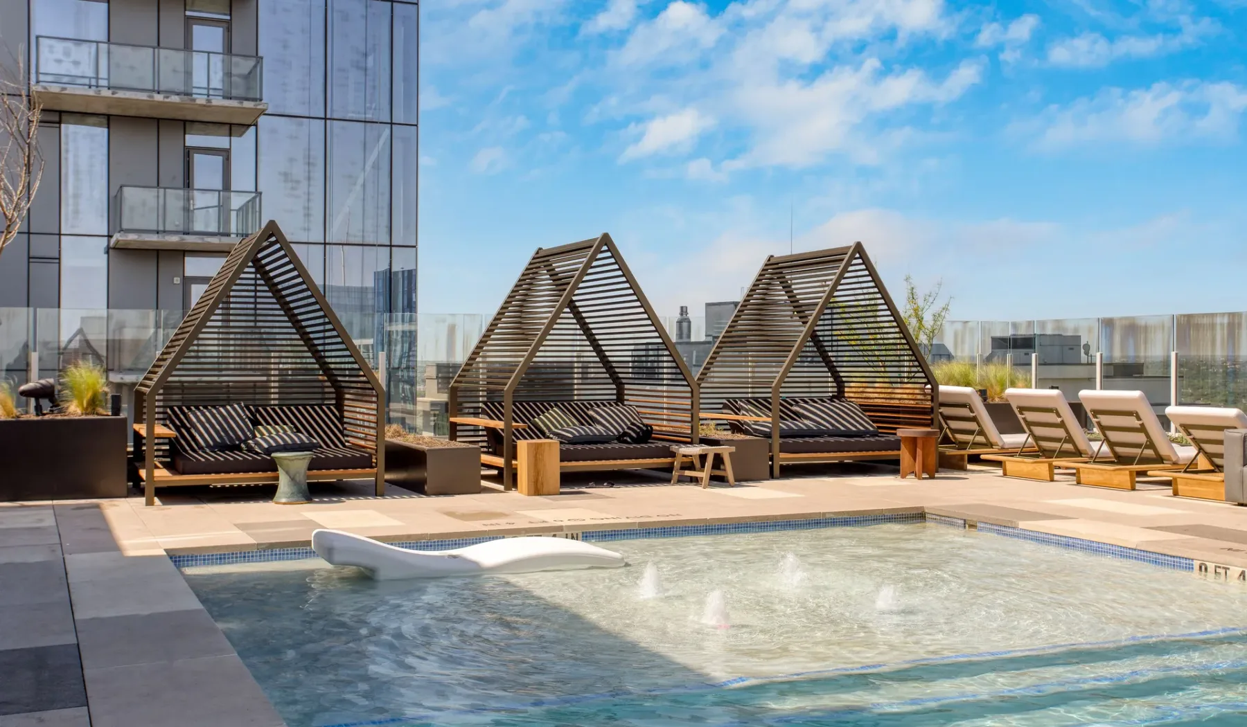 A rooftop pool with lounge seating and wooden canopy structures beside a modern building.