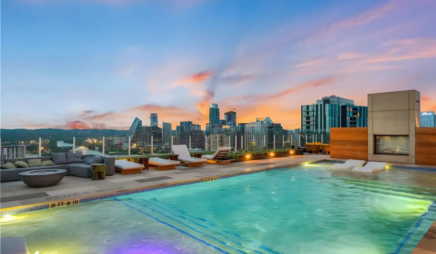 A rooftop swimming pool overlooks a city skyline at sunset.