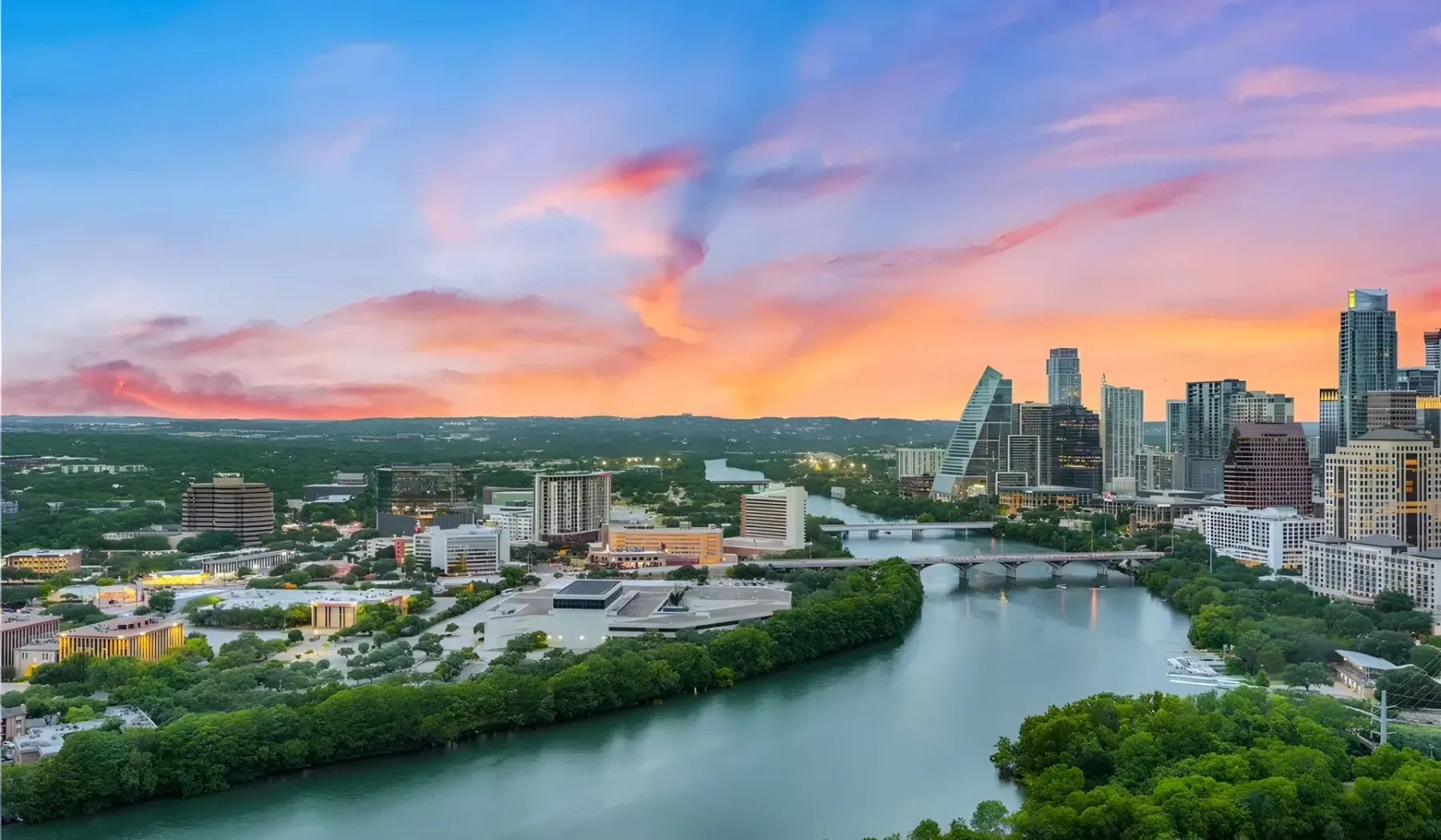 A city skyline rises beside a winding river beneath a colorful sunset sky.