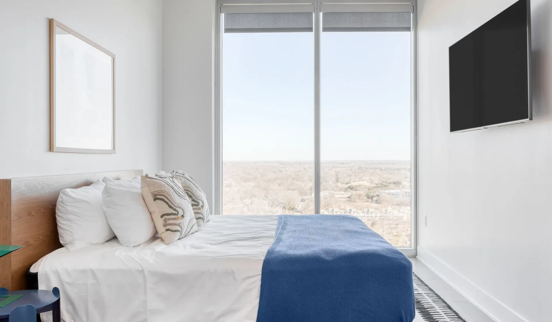 Un dormitorio luminoso con una cama, una manta azul y grandes ventanas con vistas a la ciudad.