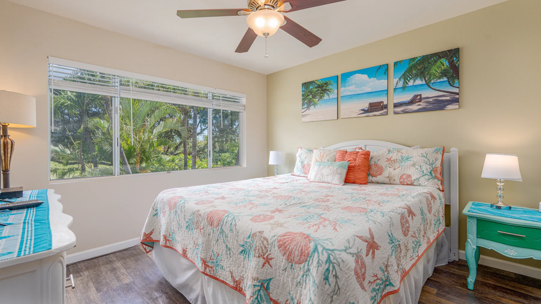 Un dormitorio con una colcha floral, grandes ventanas y arte tropical en la pared.