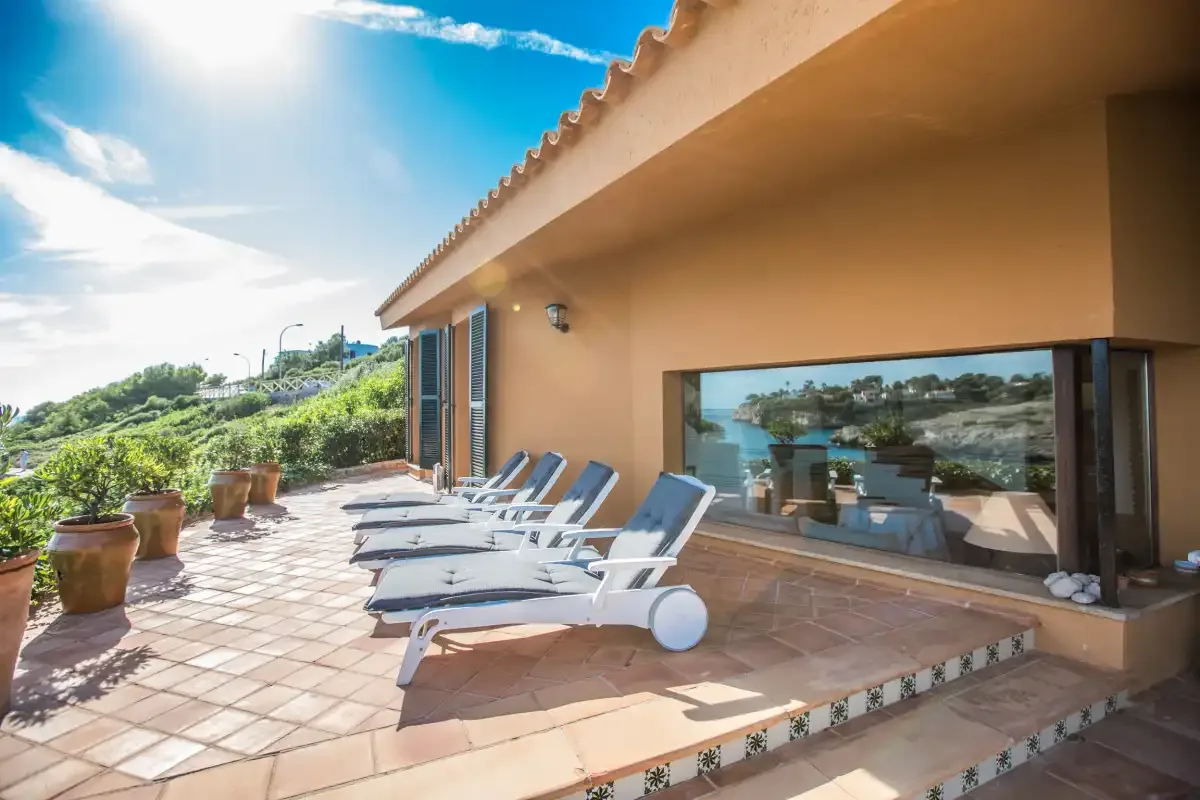 A sunny outdoor terrace beside a building has lounge seating and a large window.