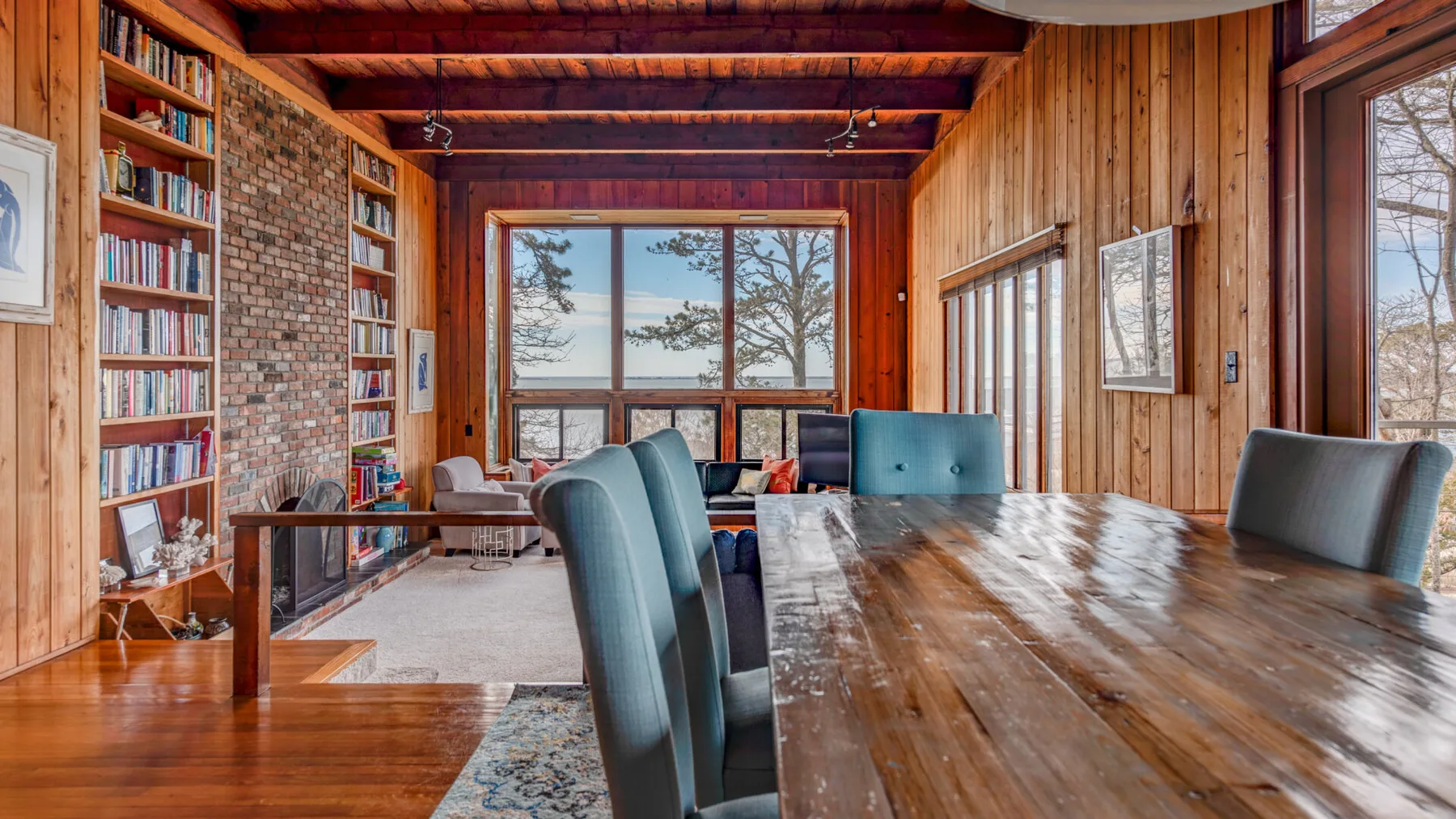 Un comedor y zona de lectura revestidos de madera, con una mesa larga, estanterías y grandes ventanas.