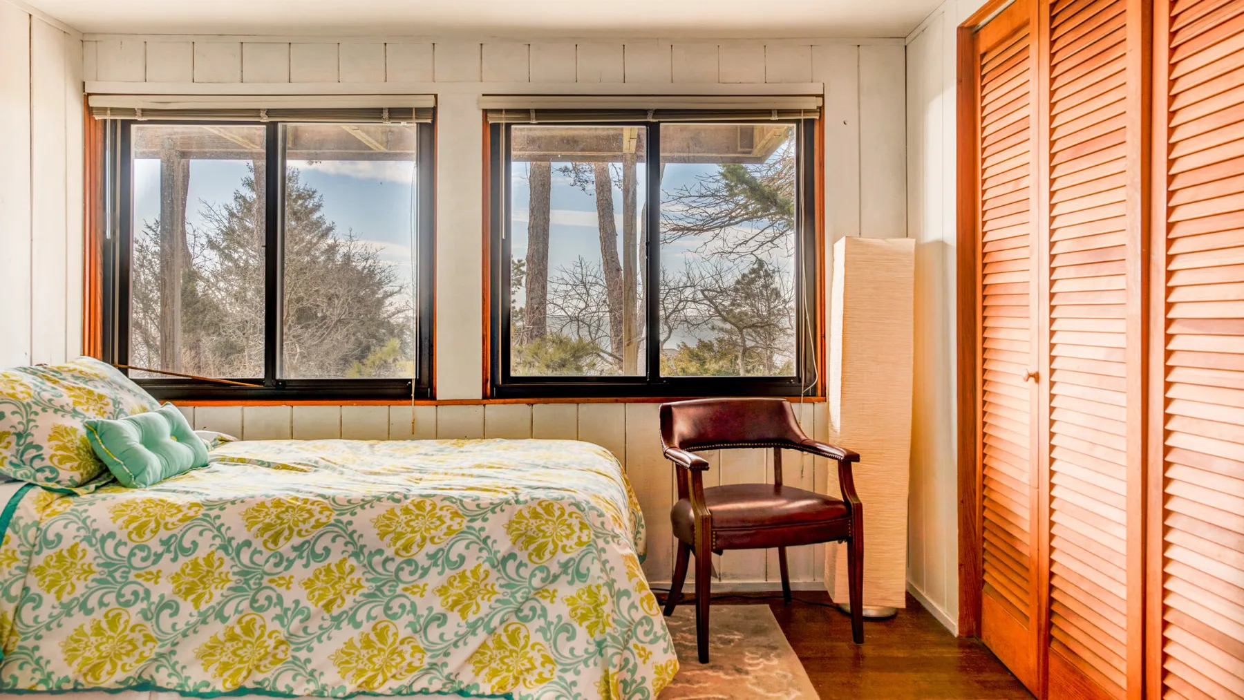 Un dormitorio con una cama estampada, dos ventanas y una silla de madera.