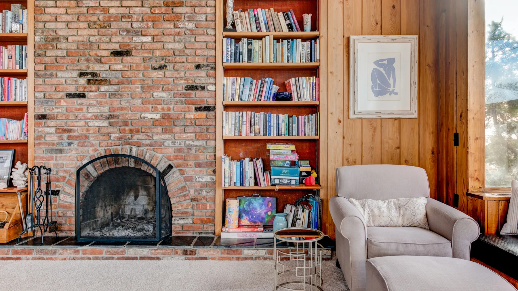Un acogedor rincón de lectura tiene una chimenea de ladrillo, estanterías y un sillón tapizado.