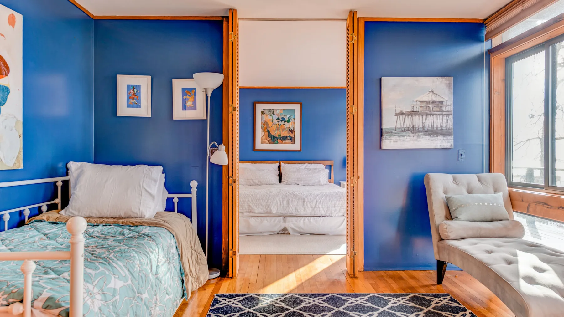 Un dormitorio azul con ropa de cama blanca, cuadros enmarcados y una chaise junto a la ventana.