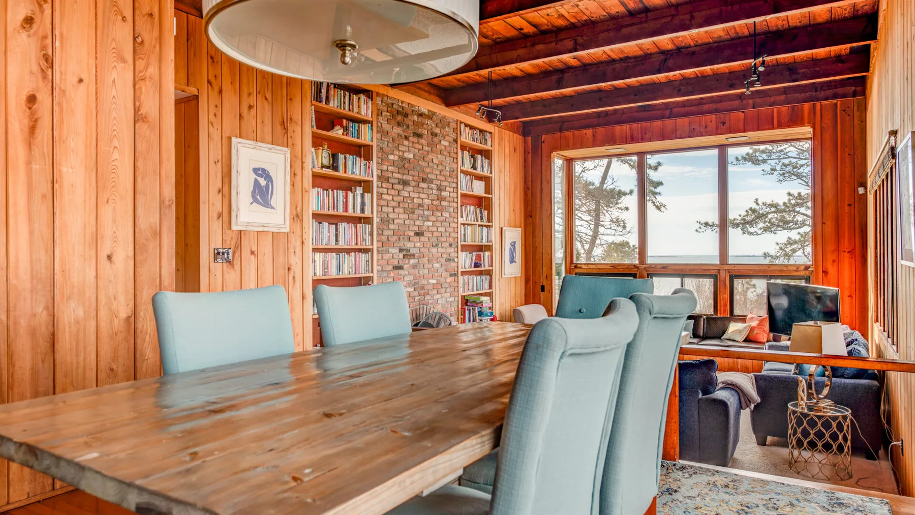 Comedor revestido de madera con una mesa larga y asientos junto a grandes ventanas.