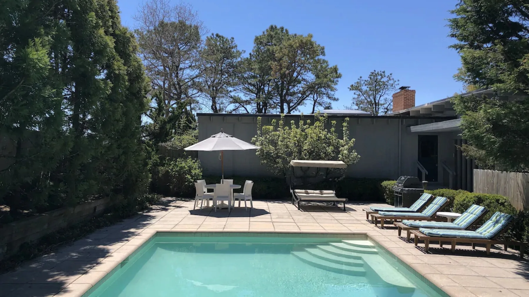 Una piscina al aire libre con asientos reclinables y una sombrilla en un patio ajardinado.