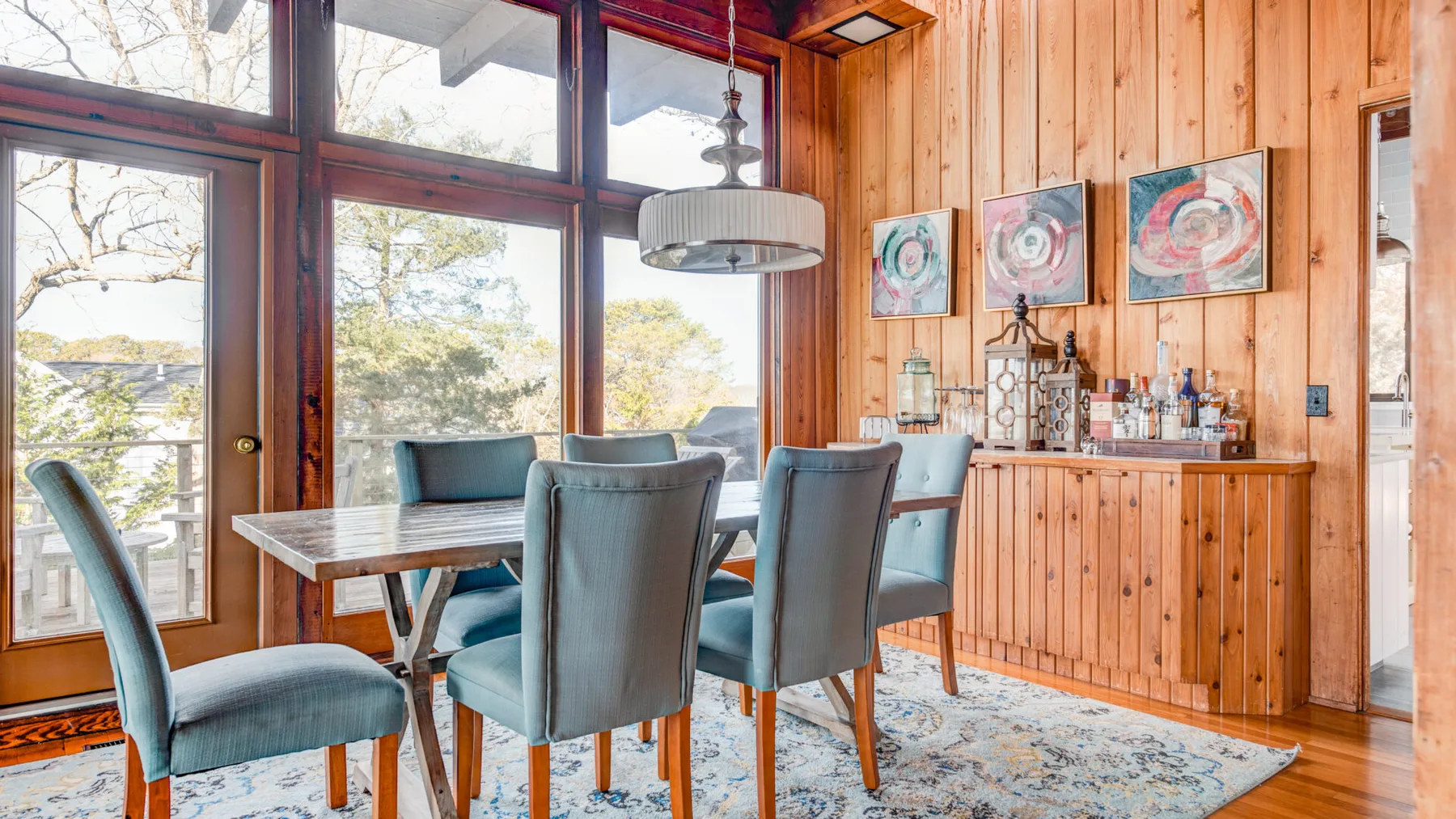 Un comedor con una mesa de madera, asientos tapizados y grandes ventanas.