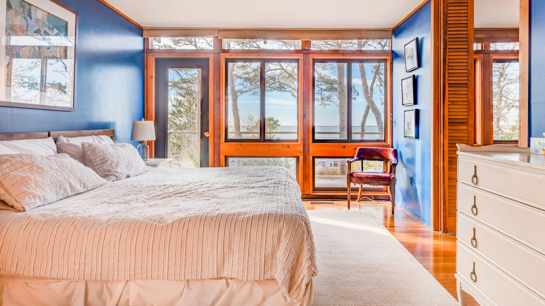 Un dormitorio con una cama grande, paredes azules y ventanas con vistas a los árboles.