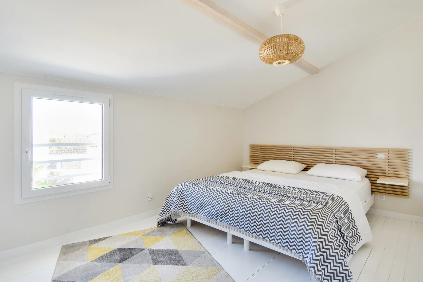 A bright bedroom with a bed covered by a patterned blanket and a window.