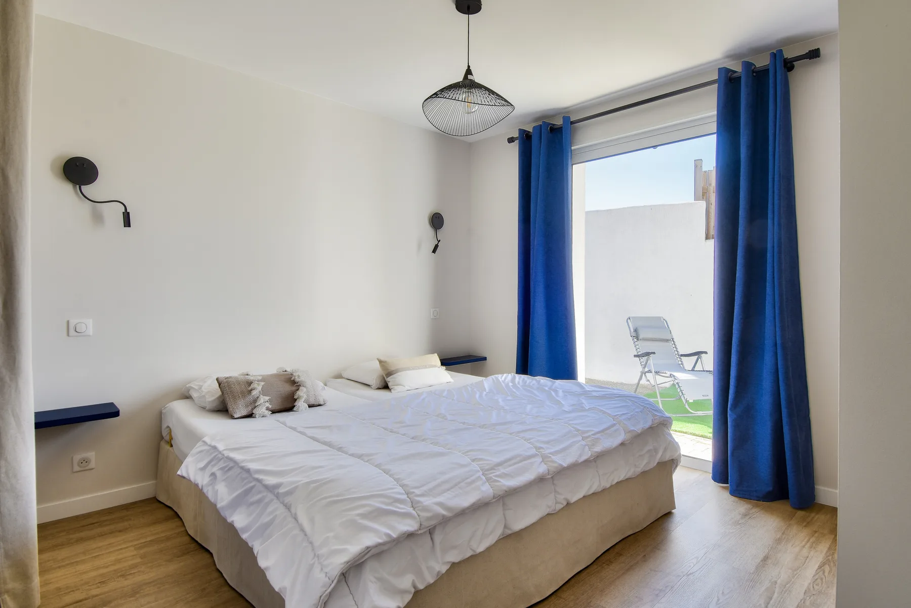 A bedroom with a bed beside blue curtains opening onto a small outdoor area.