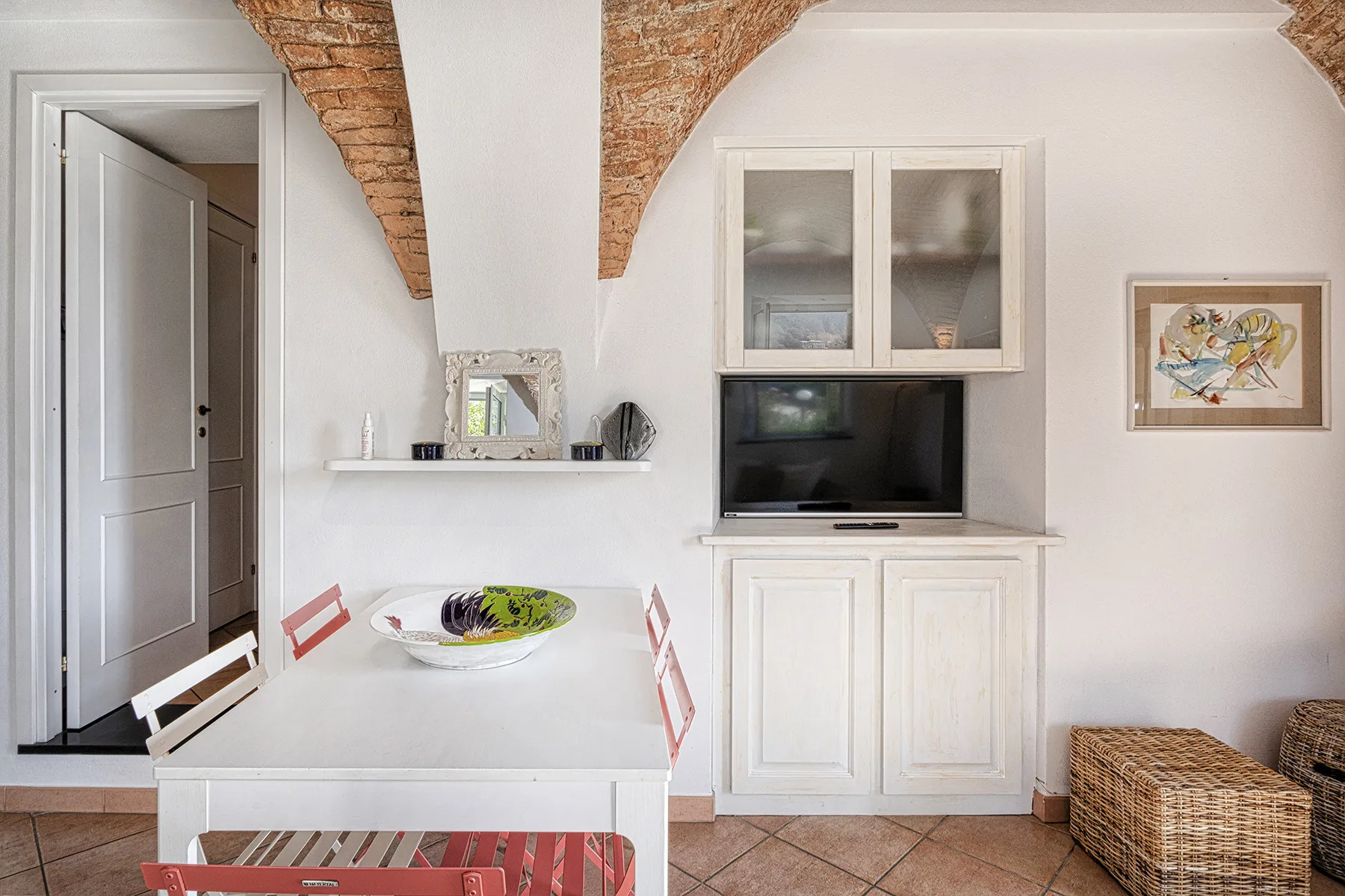 A bright dining area with a white table, television, and exposed brick ceiling beams.