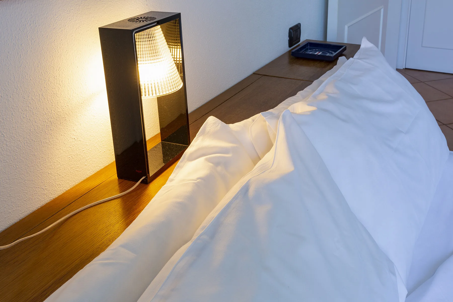 A bed with white bedding sits beside a glowing bedside lamp.