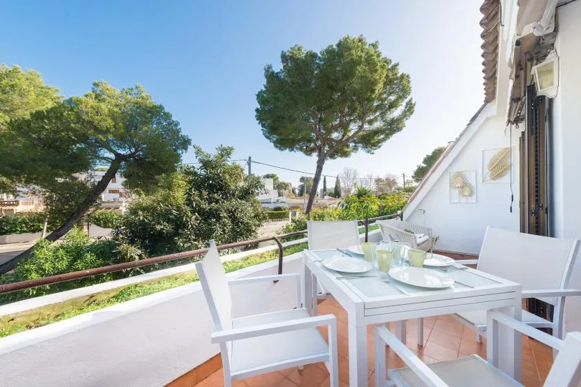 An outdoor dining area with a table and white seating overlooks leafy trees.