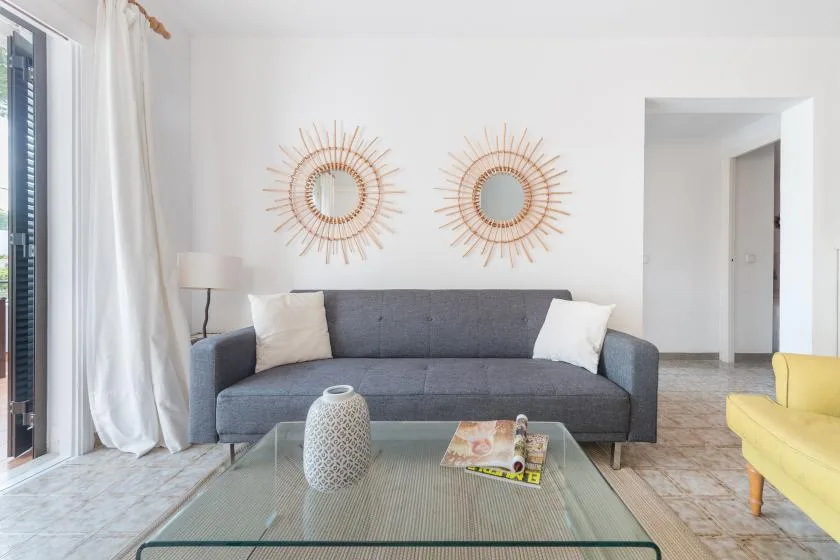 Una sala luminosa con sofá gris, mesa de centro de vidrio y espejos en la pared.