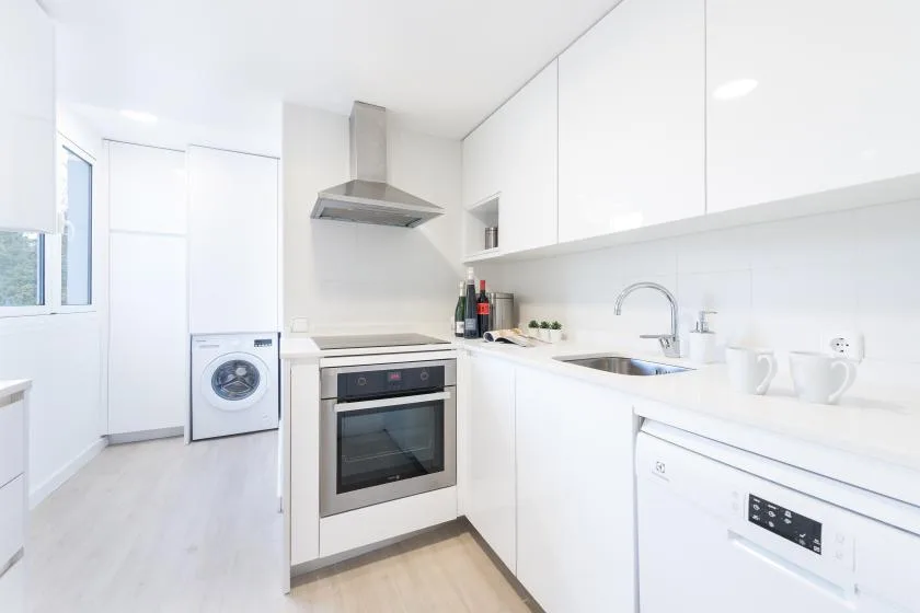 Una cocina blanca y luminosa con horno, placa de cocina, fregadero y lavadora.