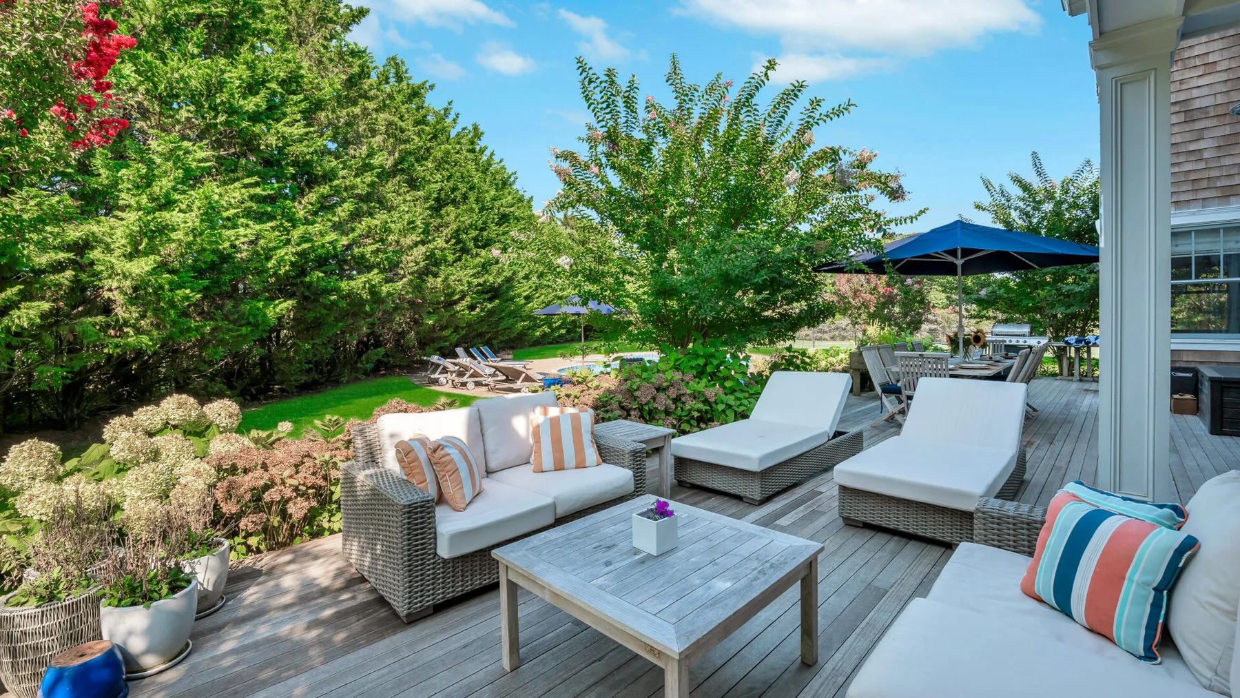 An outdoor deck features cushioned seating and a table beside a leafy garden.