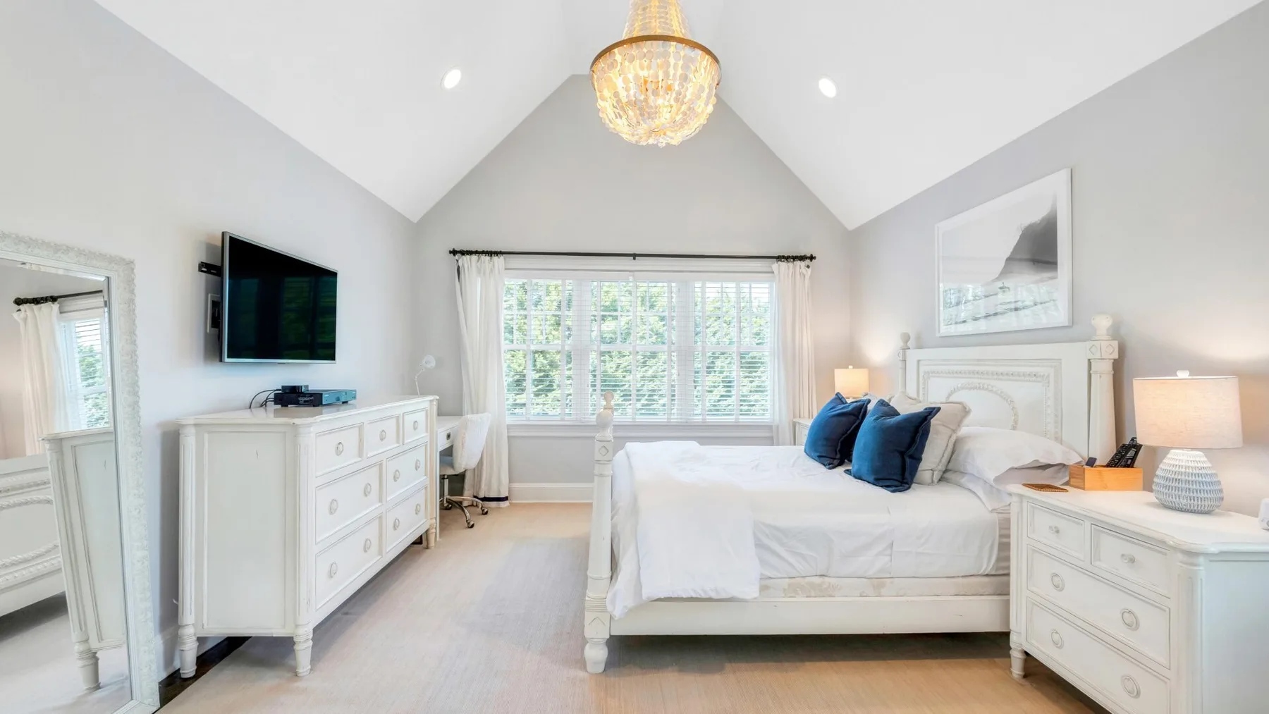 A bright bedroom with a bed beneath a large window and a chandelier overhead.