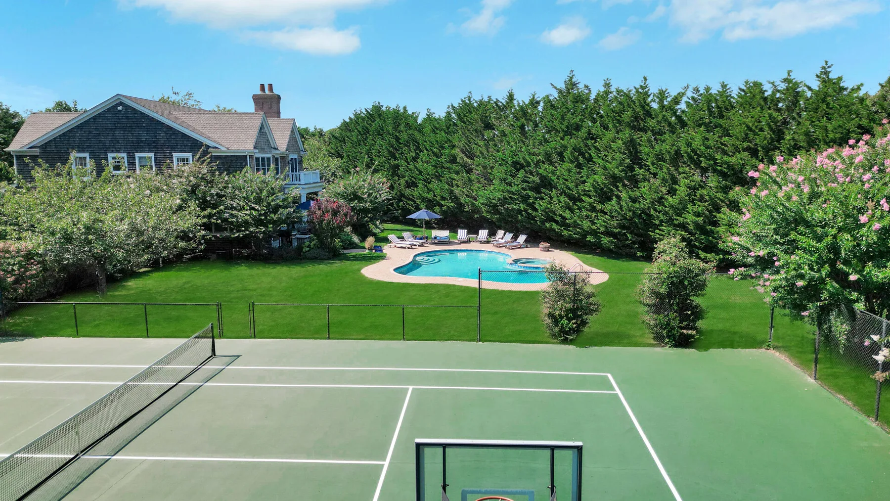 An outdoor tennis court overlooks a swimming pool and landscaped garden.