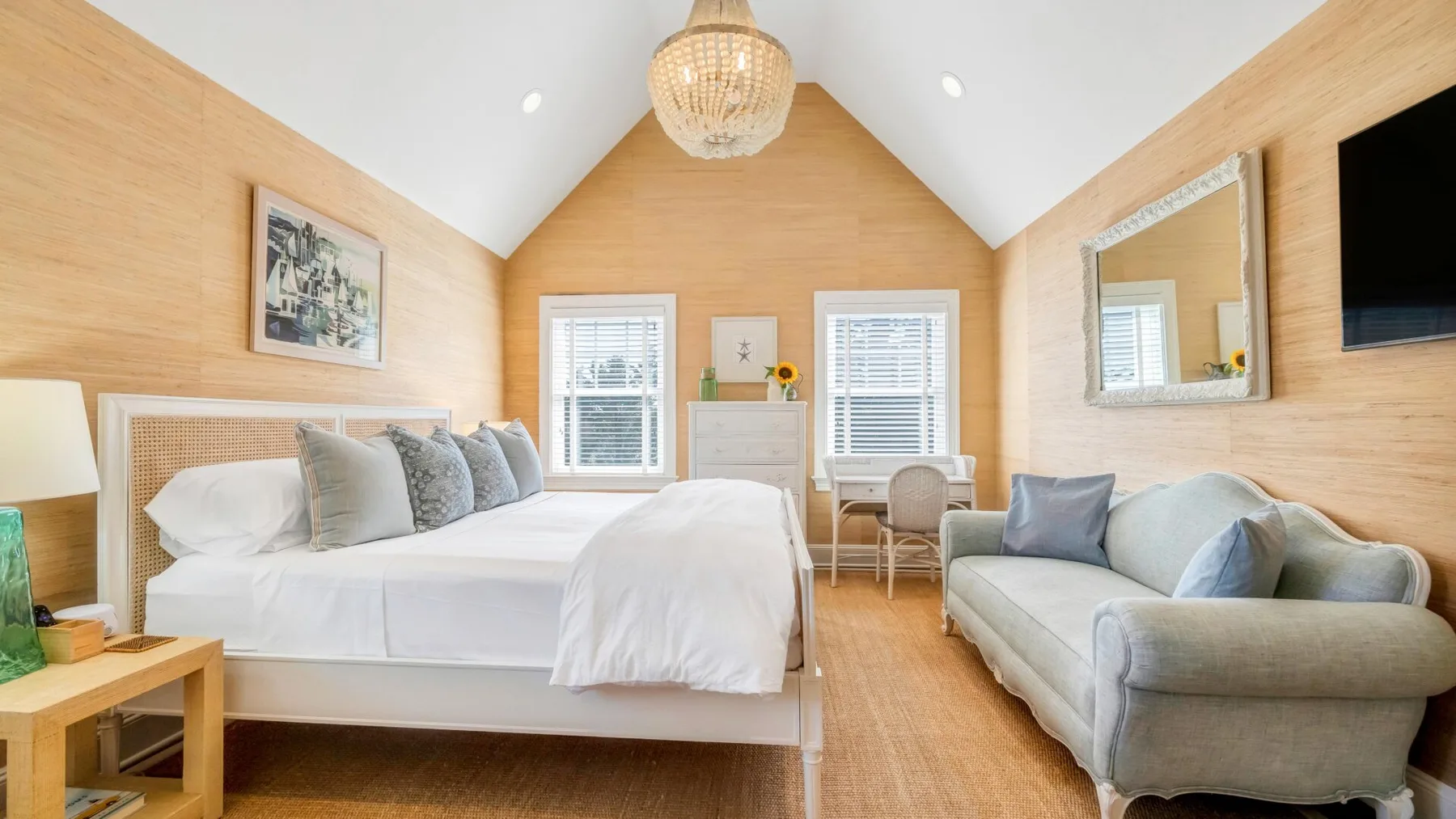 A bright bedroom with a bed, a seating area, and a desk by the windows.