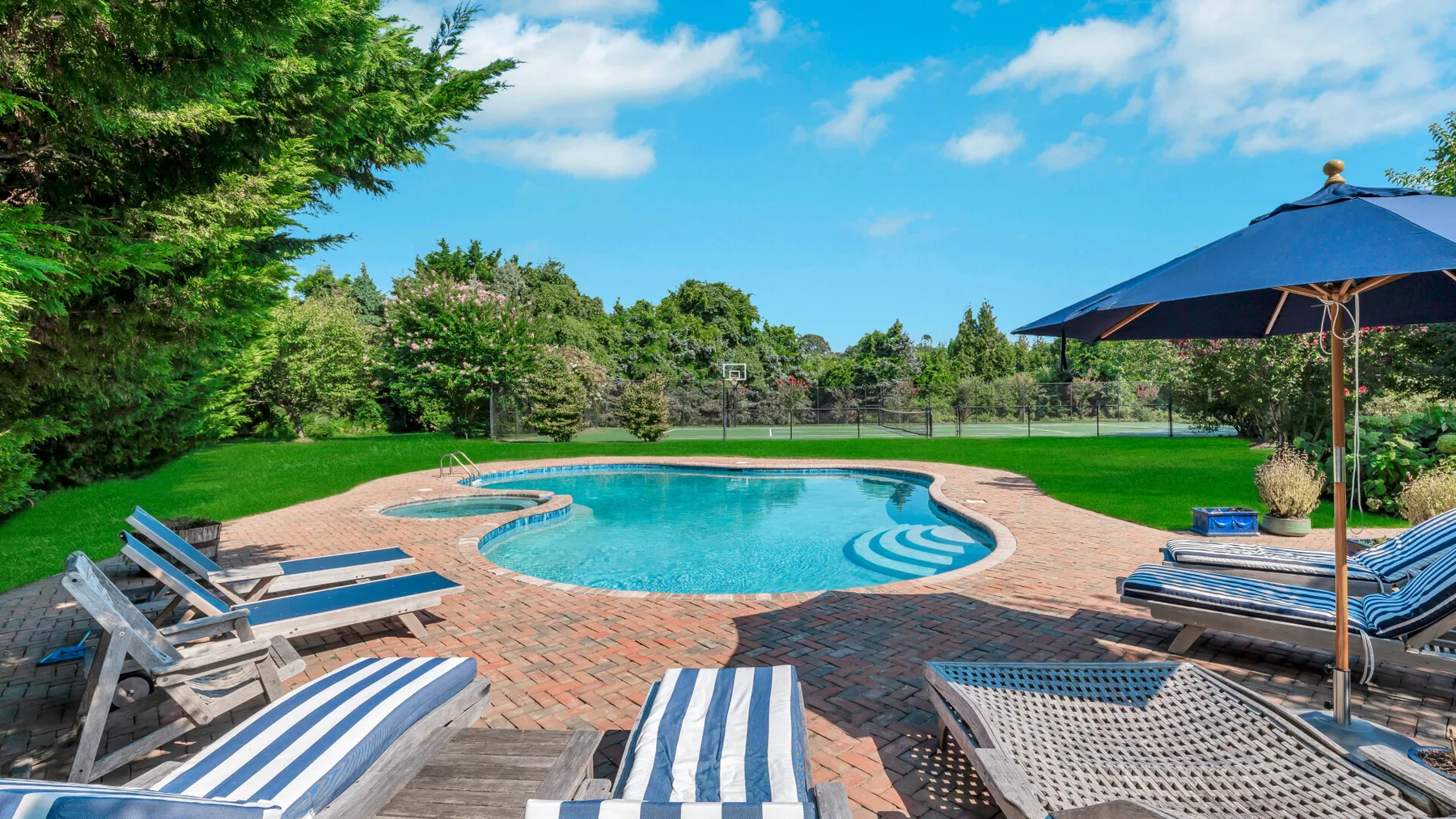 A swimming pool sits in a brick patio with striped loungers and a blue umbrella.