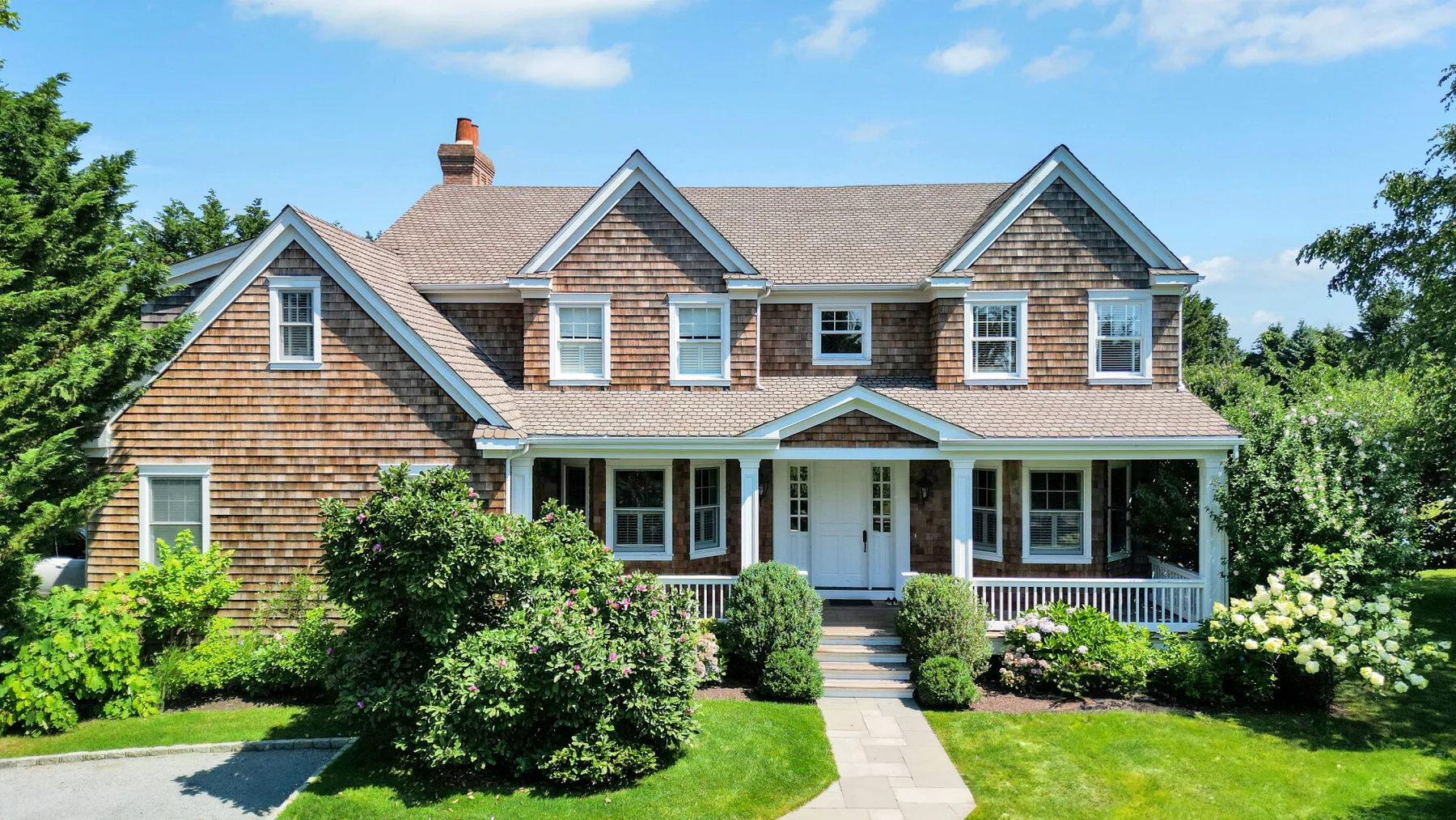 A large house with a covered porch stands behind a green lawn and flowering shrubs.