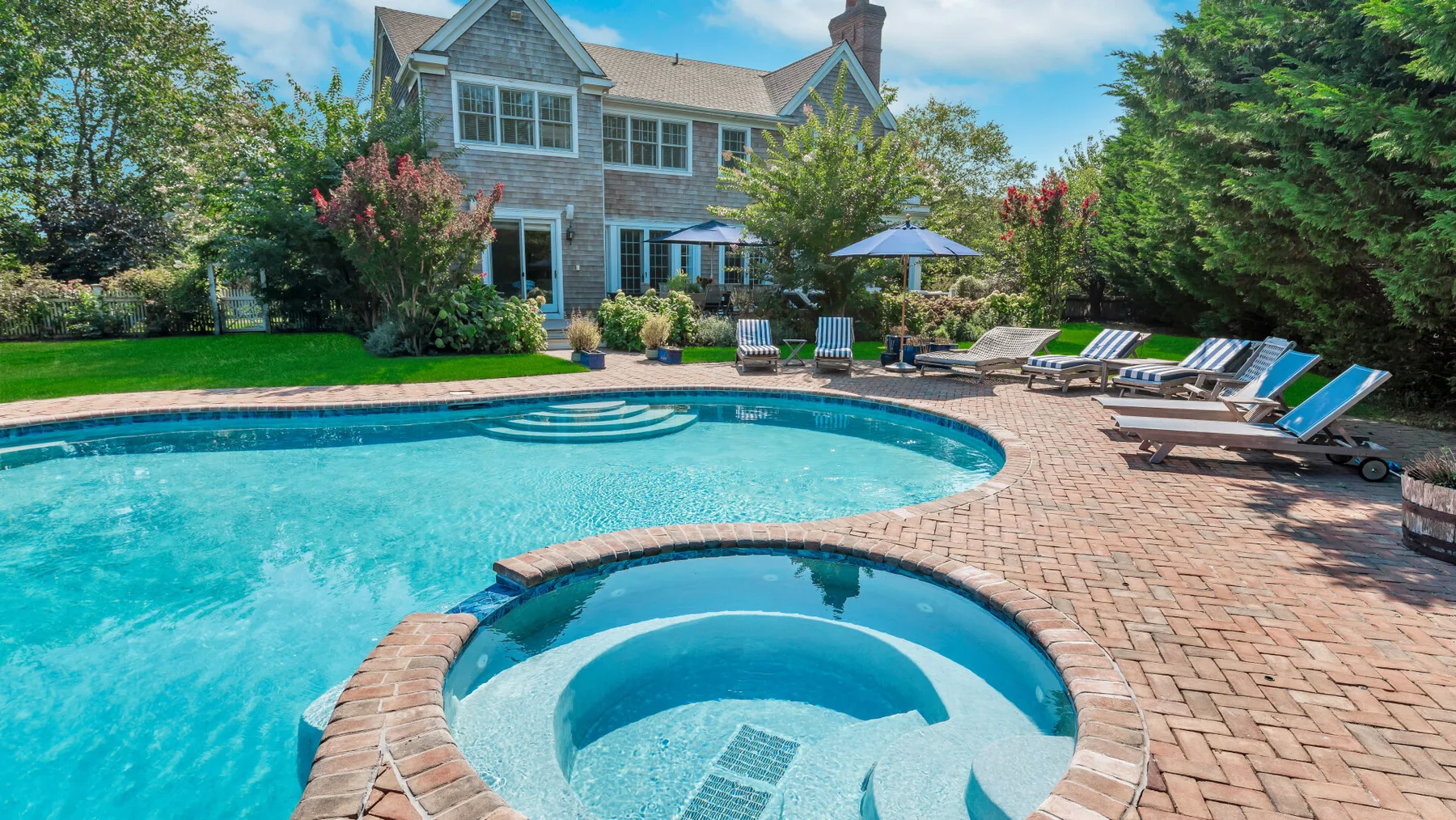 A swimming pool and circular spa sit on a brick patio beside a house.