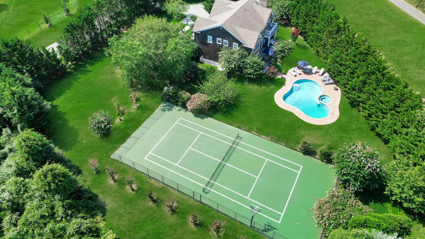 Aerial view of a tennis court, swimming pool, and house surrounded by greenery.