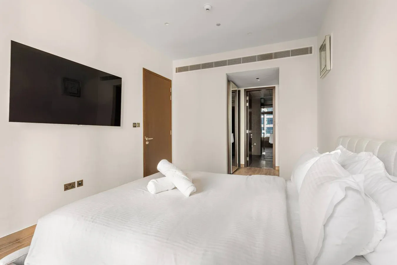 Un dormitorio luminoso con una cama, un televisor en la pared y una puerta hacia un pasillo.
