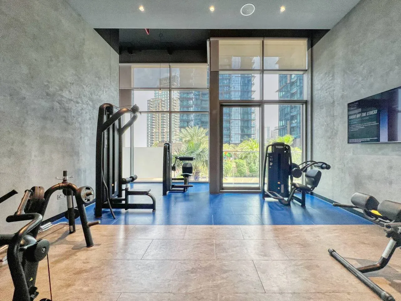 Un gimnasio con máquinas de ejercicio y grandes ventanales con edificios a la vista.