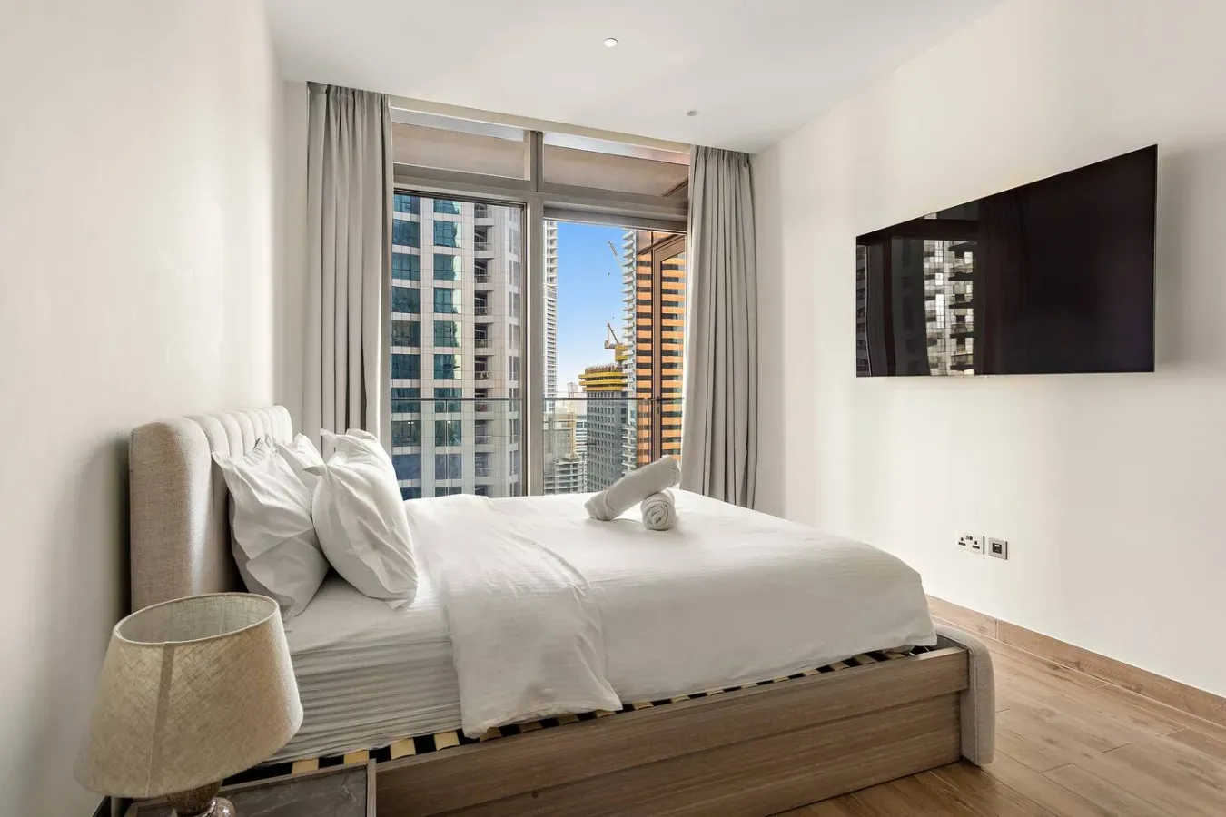 Un dormitorio con una cama bien hecha junto a una gran ventana con vistas a edificios cercanos.