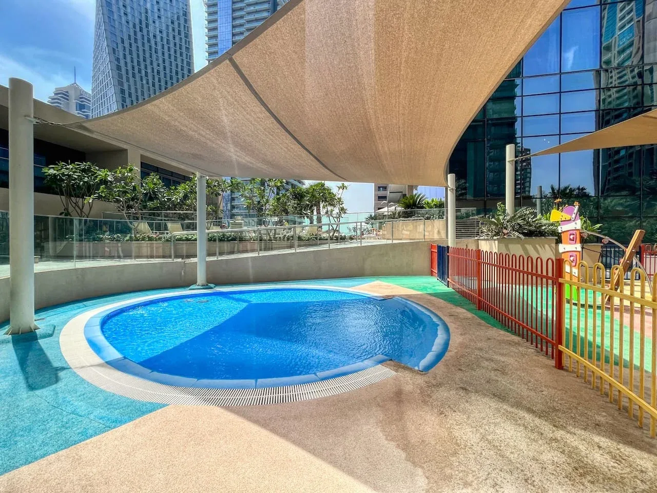Una piscina al aire libre está bajo una cubierta de sombra junto a una zona de juegos cercada.