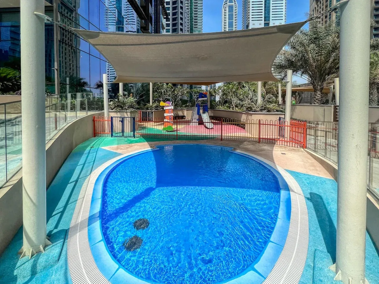 Una piscina al aire libre está bajo una cubierta de sombra, con un área de juegos detrás.