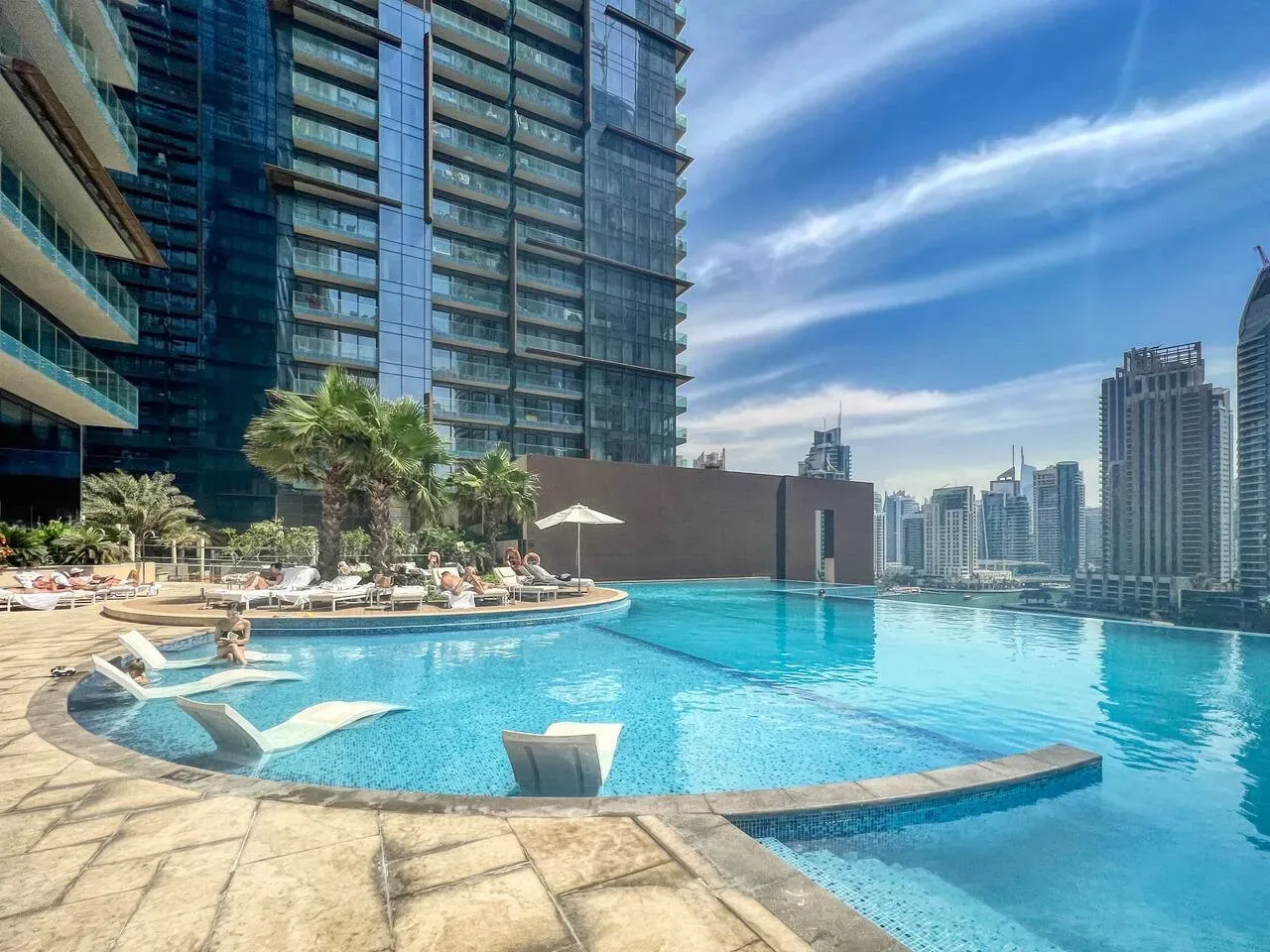 Una piscina al aire libre junto a un edificio alto moderno, con torres de la ciudad al fondo.