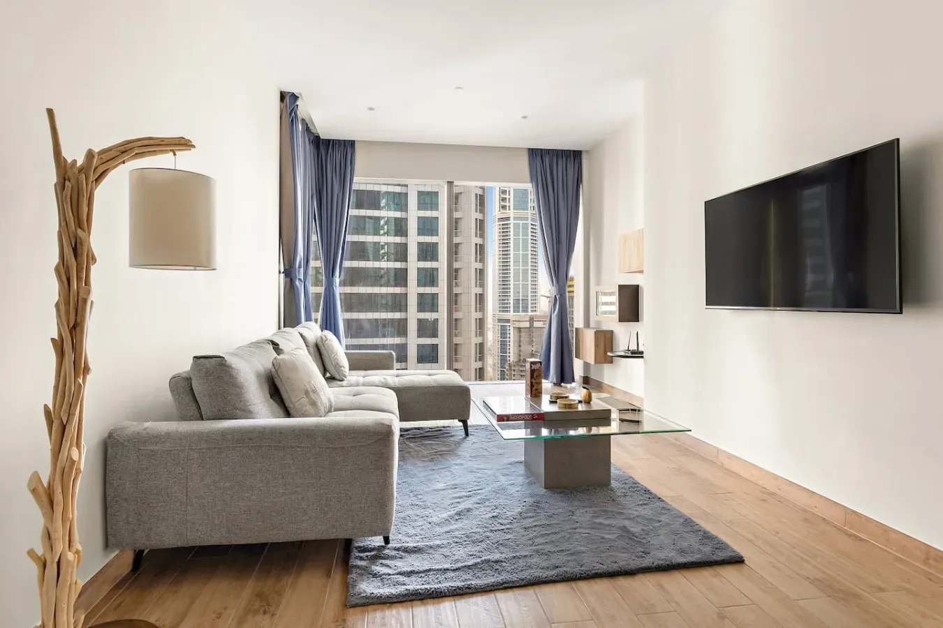 Una sala de estar luminosa con asientos grises, un televisor en la pared y grandes ventanas.