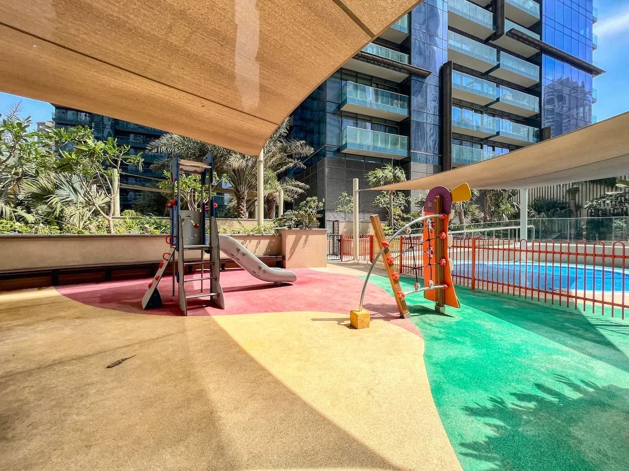 Un parque infantil al aire libre con sombra junto a una piscina, con juegos y edificios alrededor.