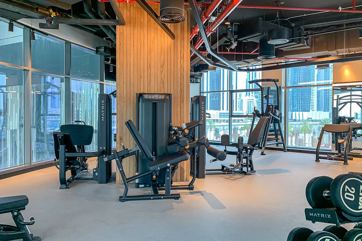 Un gimnasio moderno con máquinas de ejercicio junto a grandes ventanas con vistas a edificios de la ciudad.