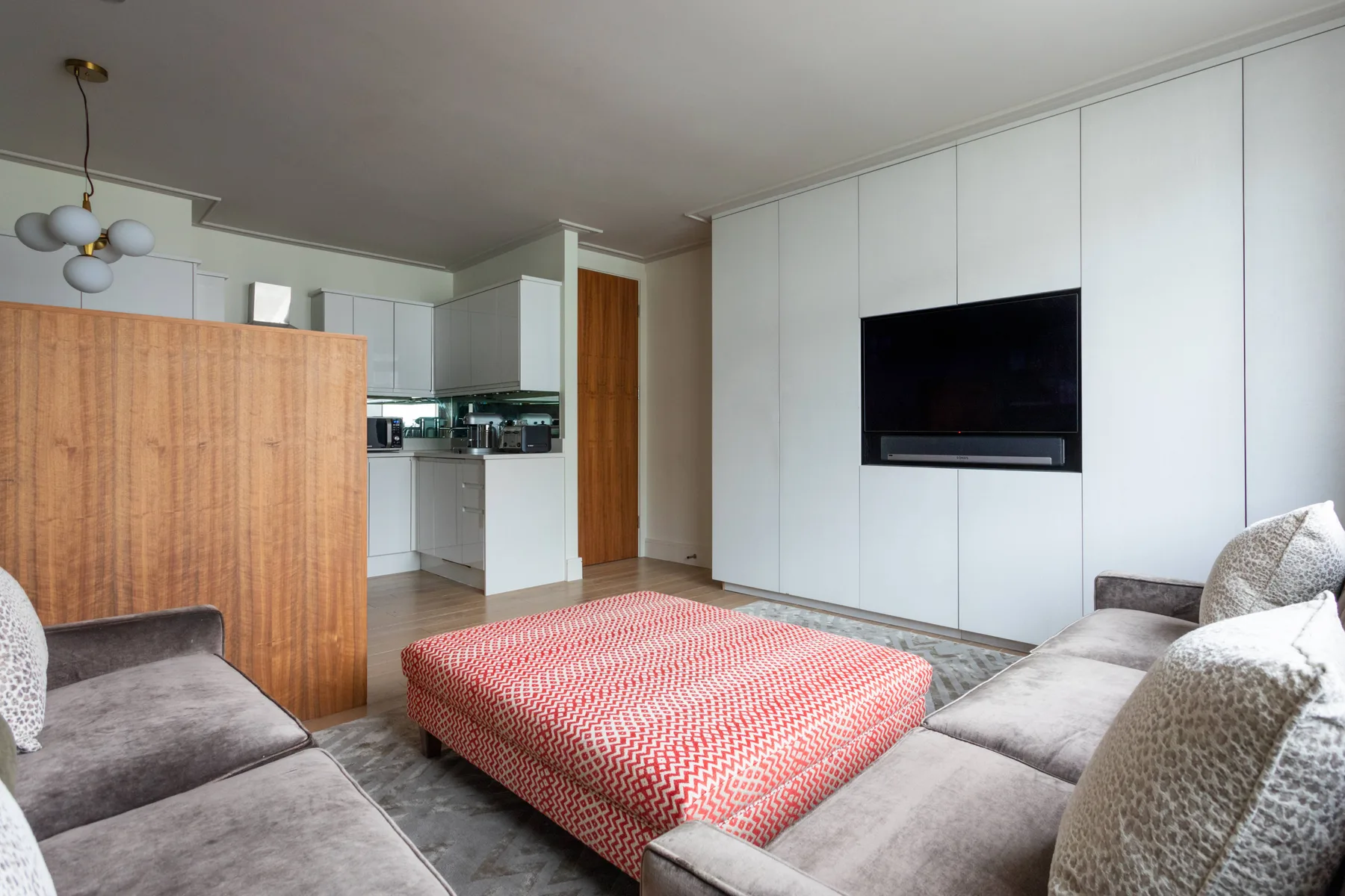 A living room with seating around a red patterned ottoman and a wall-mounted television.
