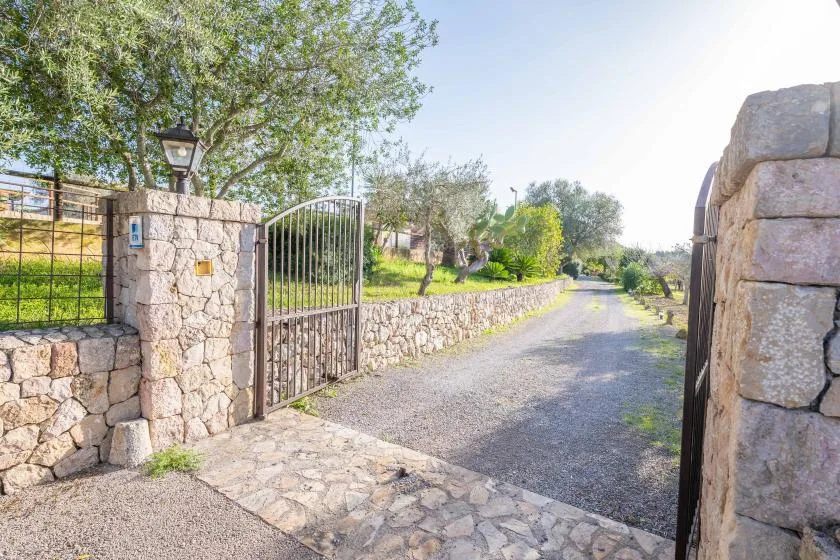 Una puerta metálica abierta da a un camino de grava bordeado por muros de piedra y árboles.