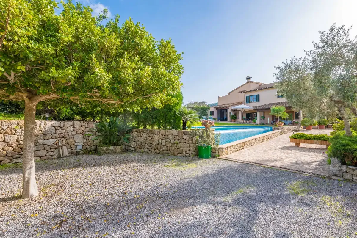 Un patio de grava conduce a una piscina y una casa rodeada de árboles.