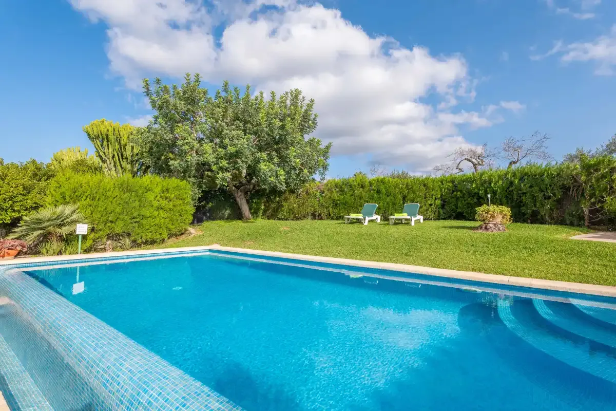 Una piscina junto a un césped, setos y árboles bajo un cielo parcialmente nublado.
