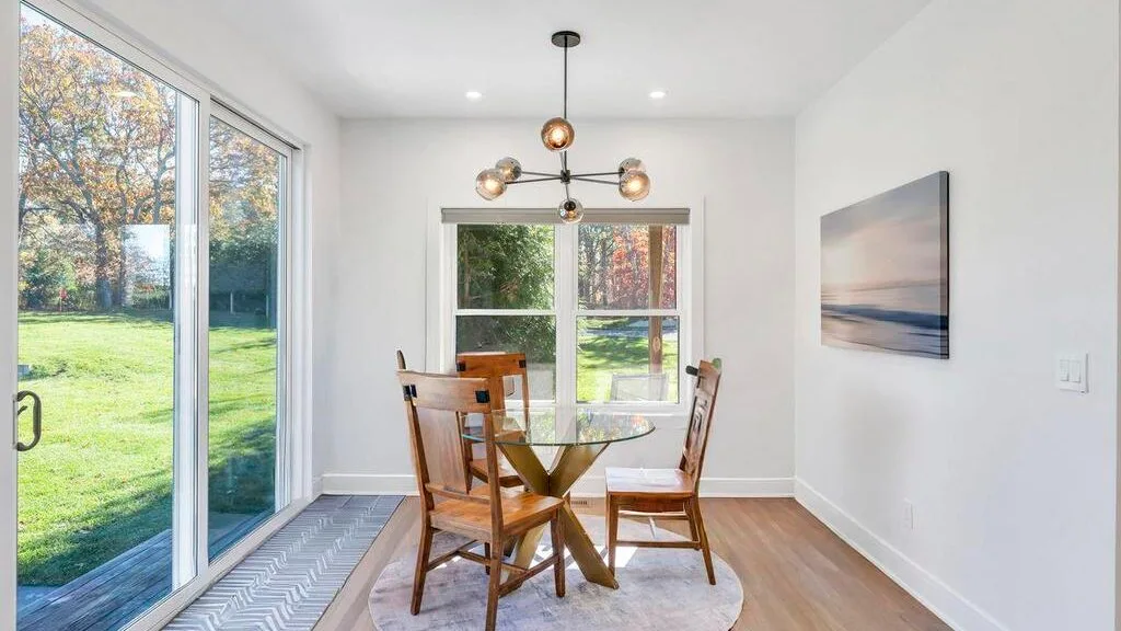 Un comedor luminoso tiene una mesa de madera y sillas junto a grandes ventanas con vistas a zonas verdes.